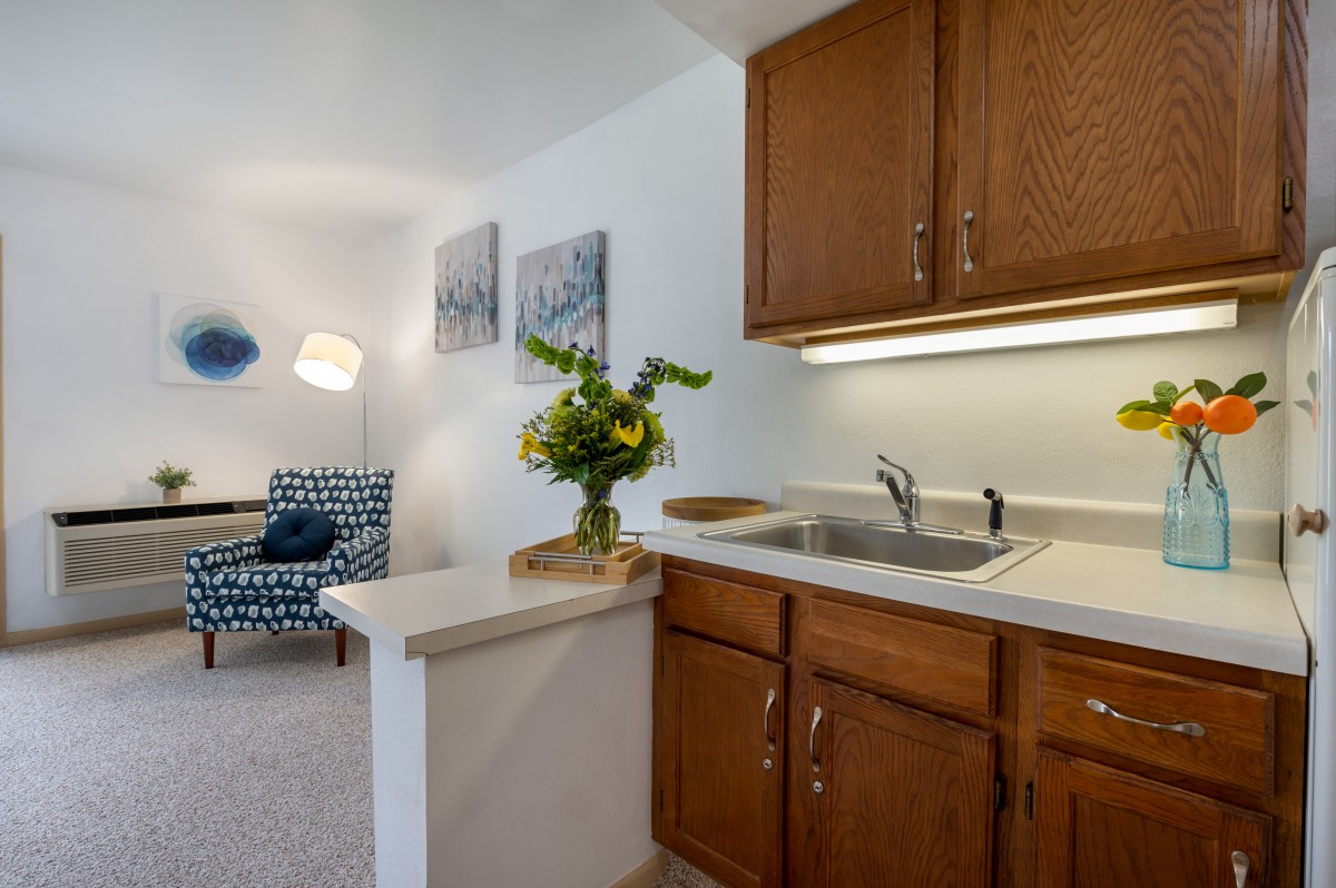 A cozy kitchen area with wooden cabinets, a sink, and decorative elements like flowers and artwork on the wall.