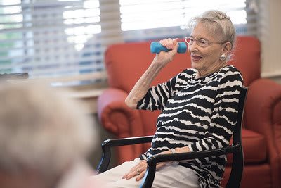 An elderly woman exercises with a dumbbell while seated indoors, smiling and wearing a striped shirt.