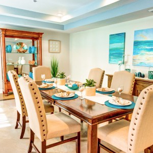A beautifully decorated dining room featuring a wooden table set with teal placemats and beige upholstered chairs, complemented by coastal artwork on the walls.