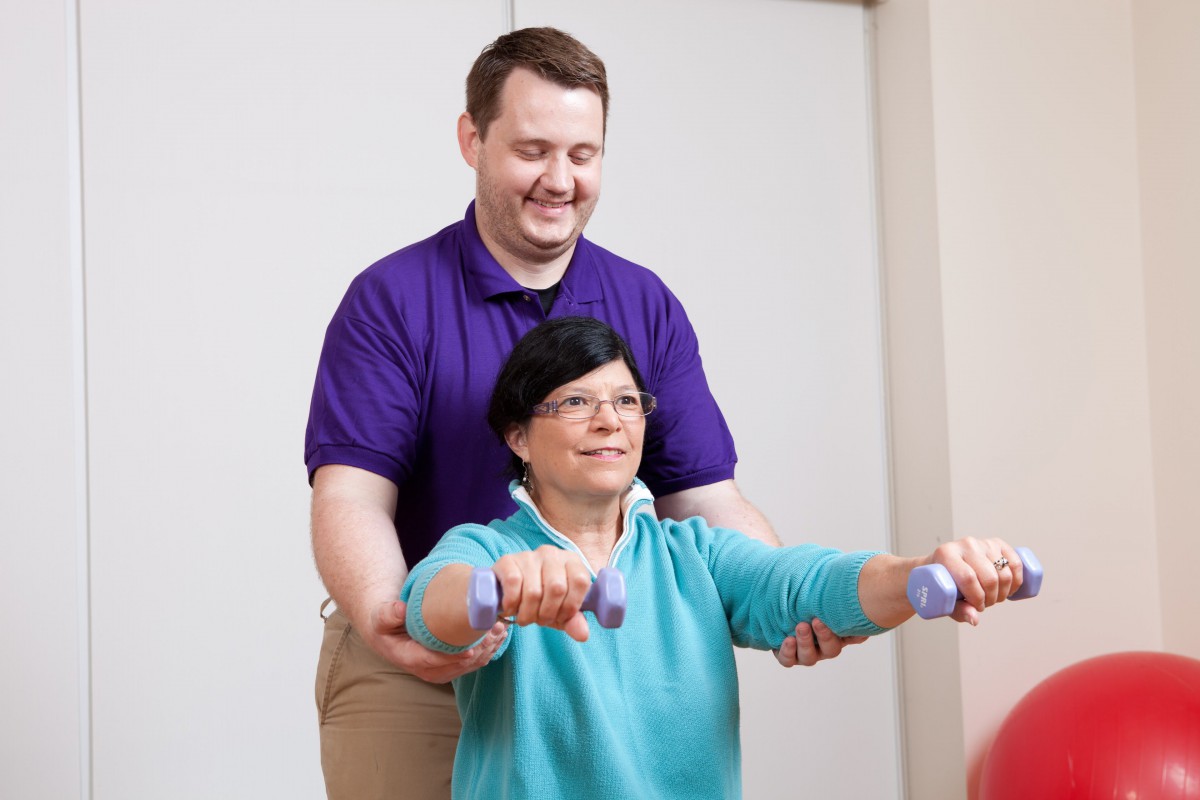 A therapist assists an elderly woman with arm exercises using dumbbells.