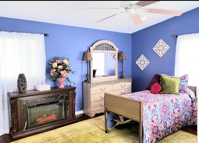A cozy bedroom with blue walls, featuring a bed with colorful bedding, a dresser with a mirror, and decorative elements like floral arrangements and a fireplace.