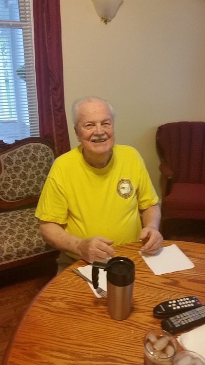 An elderly man in a yellow shirt is smiling while seated at a table with papers and a mug.