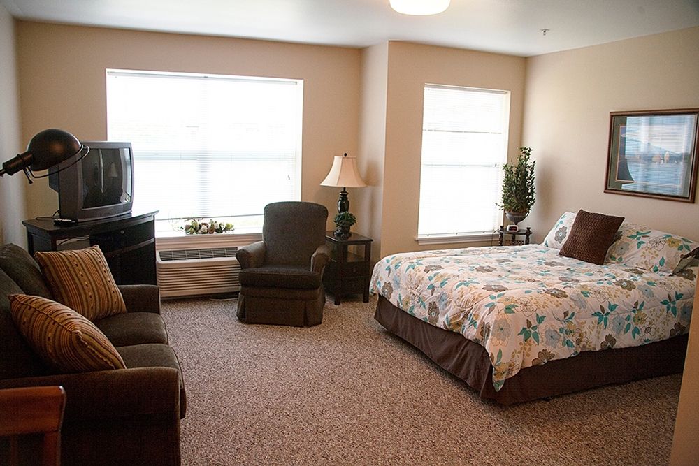 A well-furnished bedroom with two windows, featuring a bed, chairs, lamp, and television.