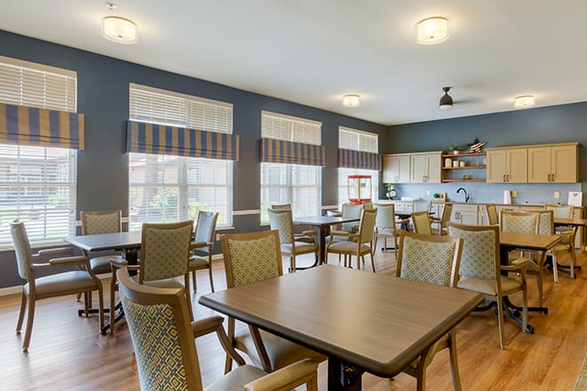 A brightly lit dining room with multiple tables and chairs, featuring large windows and an adjacent kitchen area.