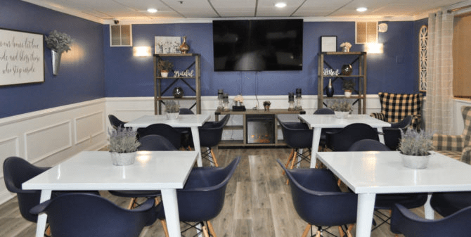 A modern dining area with sleek white tables, navy chairs, and decorative shelving under a large television.