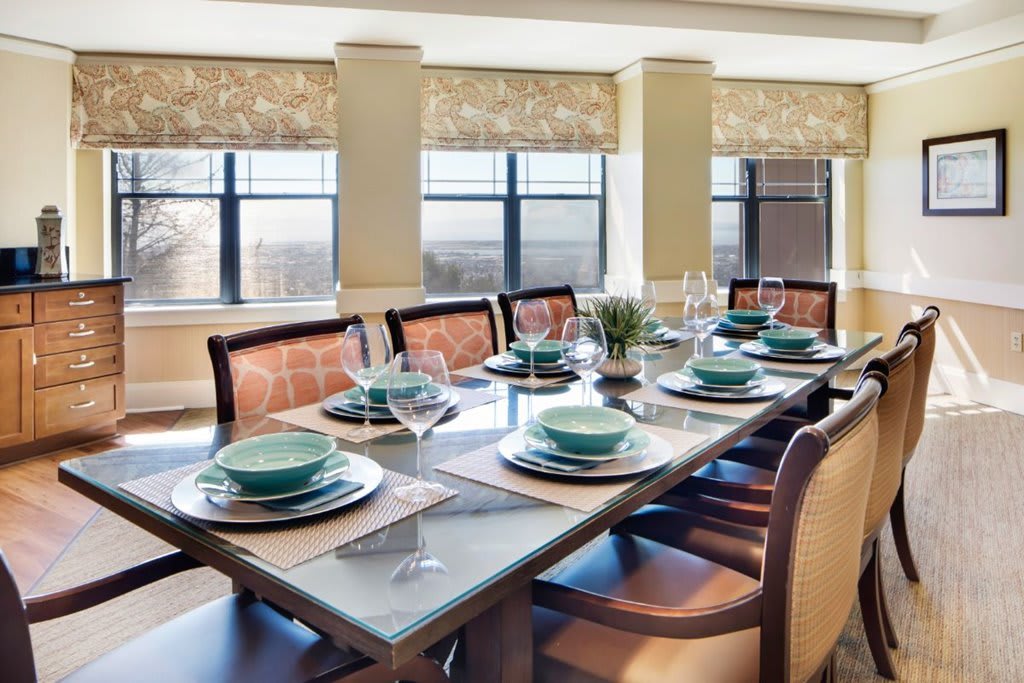 A beautifully set dining table with green dishes and wine glasses, surrounded by upholstered chairs in a bright room with large windows.