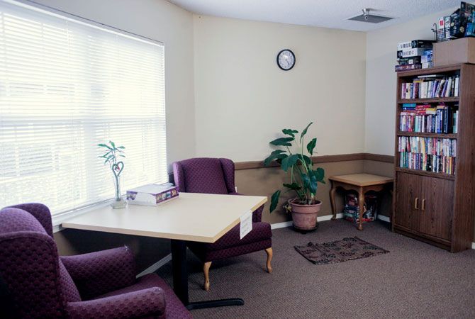 A cozy waiting room with purple chairs, a table, a potted plant, and a clock on the wall.
