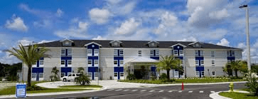 A modern blue and white hotel building with palm trees under a partly cloudy sky.