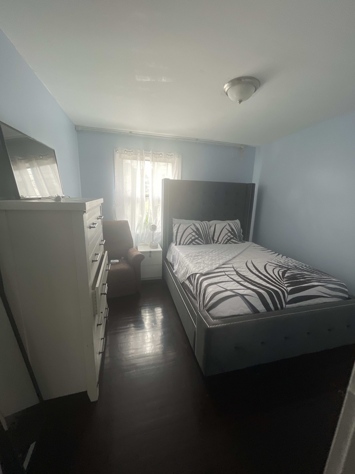 A small, cozy bedroom with a bed, dresser, chair, and a window letting in natural light.