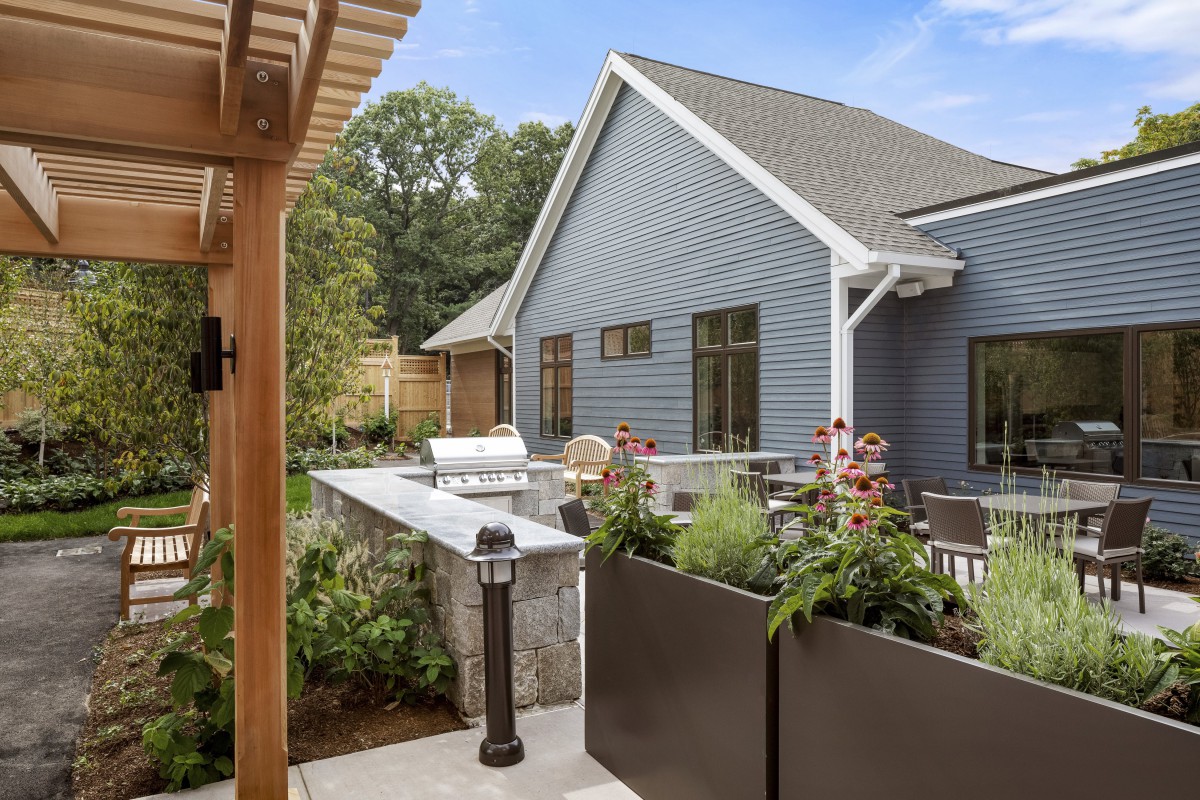 A cozy outdoor patio area featuring a barbecue grill, surrounded by lush plants and a pergola.