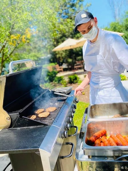 A chef grills burgers and sausages outdoors on a sunny day.
