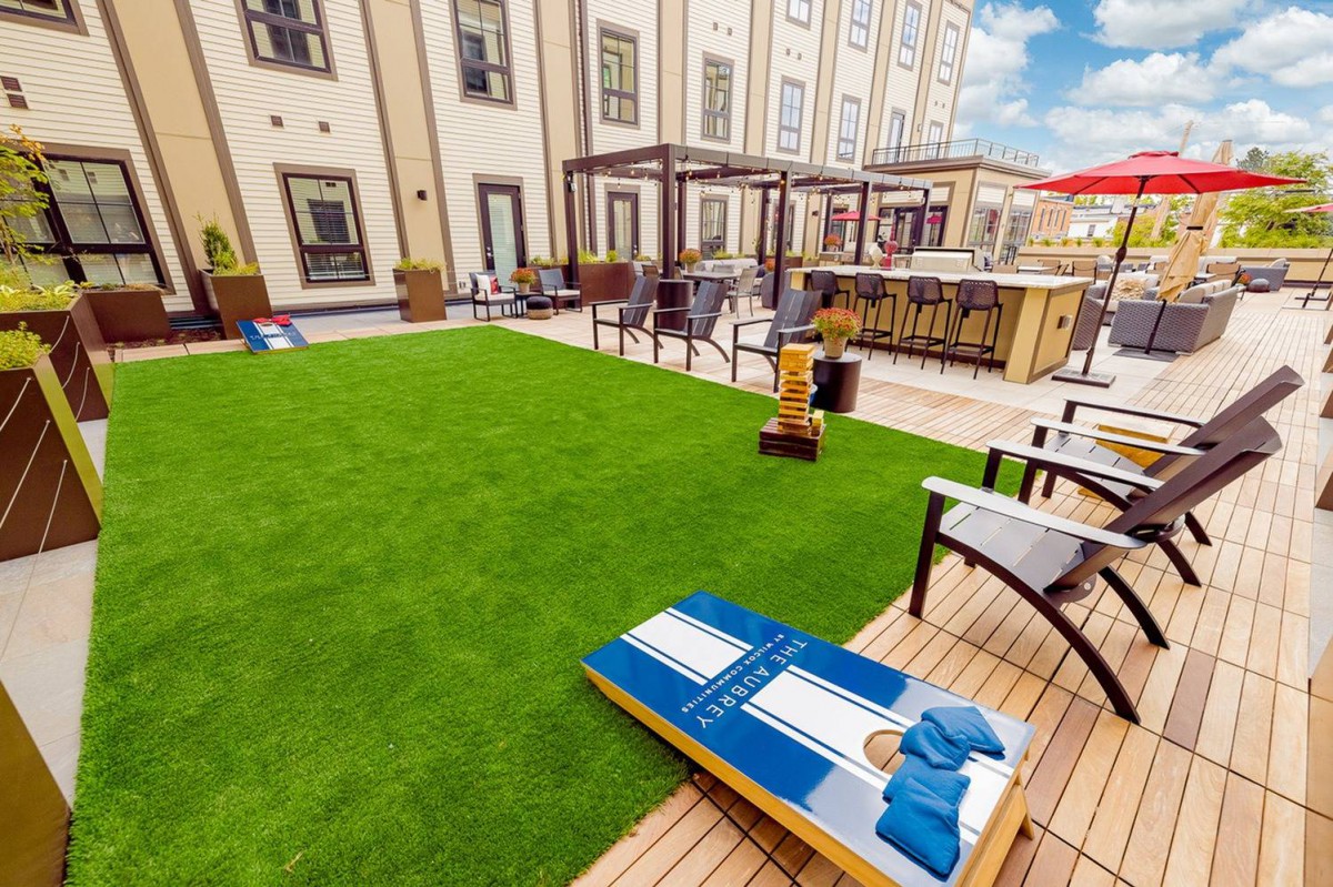 A rooftop lounge with a grass lawn, seating area, and cornhole game on a sunny day.