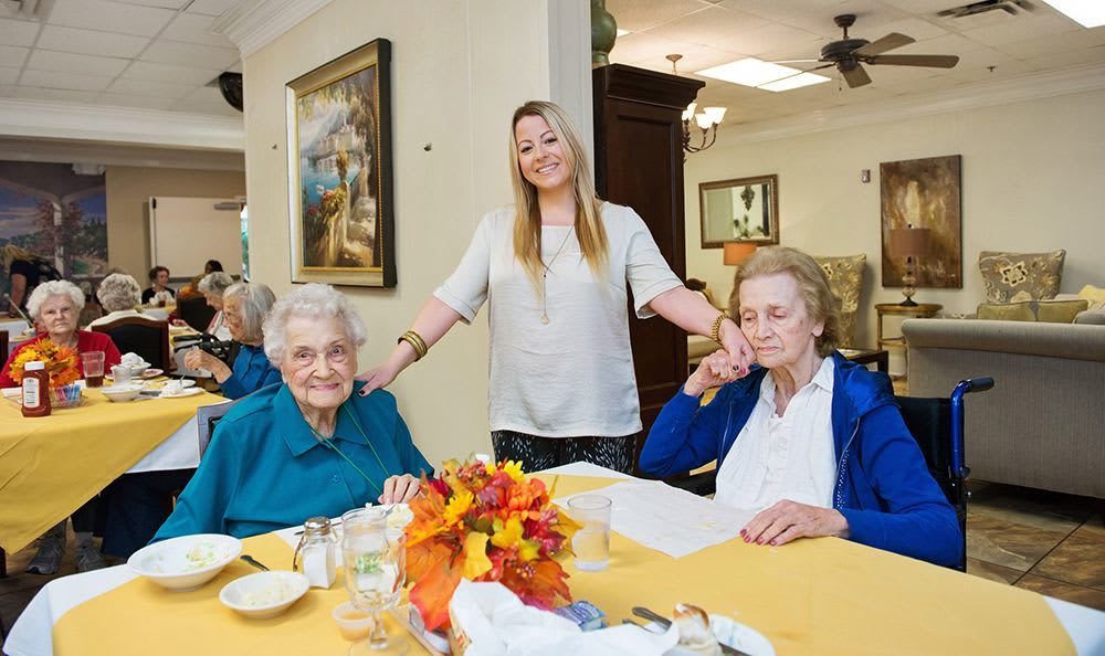 A caregiver stands smiling between two elderly women seated at a dining table, decorated with flowers.