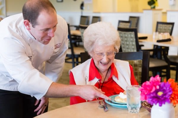 A chef is serving dessert to a smiling elderly woman in a restaurant.