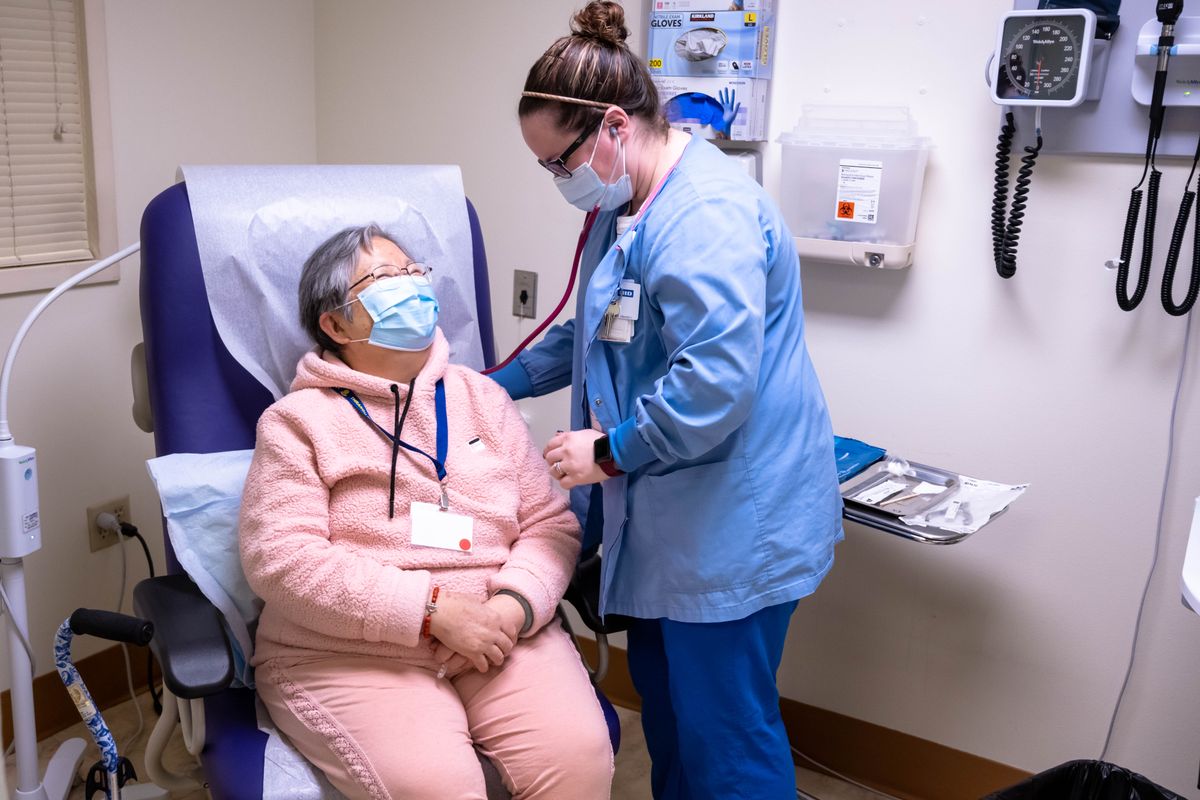 A nurse checks on a patient in a healthcare setting, both wearing masks.