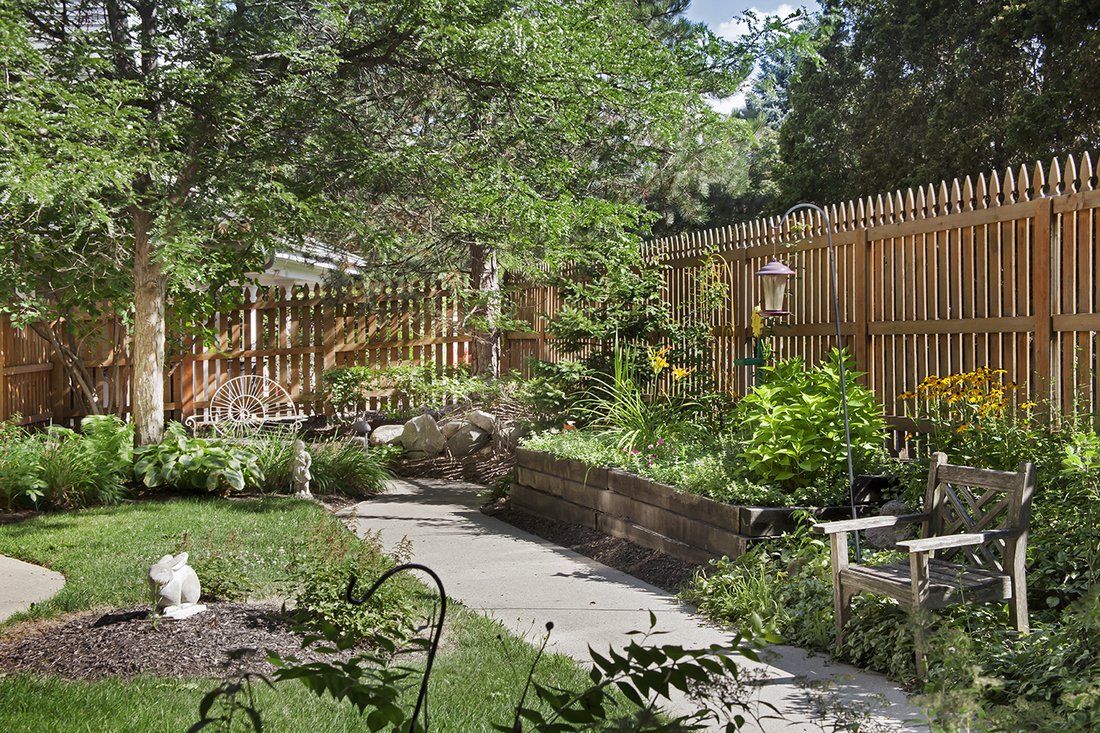A serene backyard garden featuring lush greenery, a wooden fence, and a paved pathway.