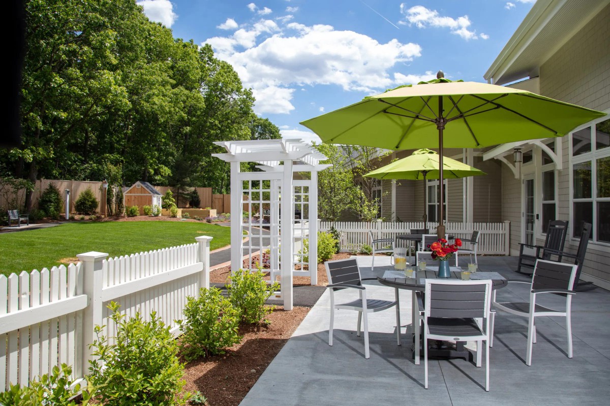 A sunny outdoor patio with a white fence, green umbrellas, and garden seating.