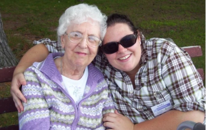 A young woman wearing sunglasses hugs an elderly woman, both smiling outdoors.