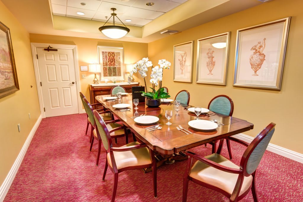 An elegantly decorated dining room with a long wooden table, surrounded by chairs and set with plates. The room features warm lighting, artwork on the walls, and a plush red carpet.