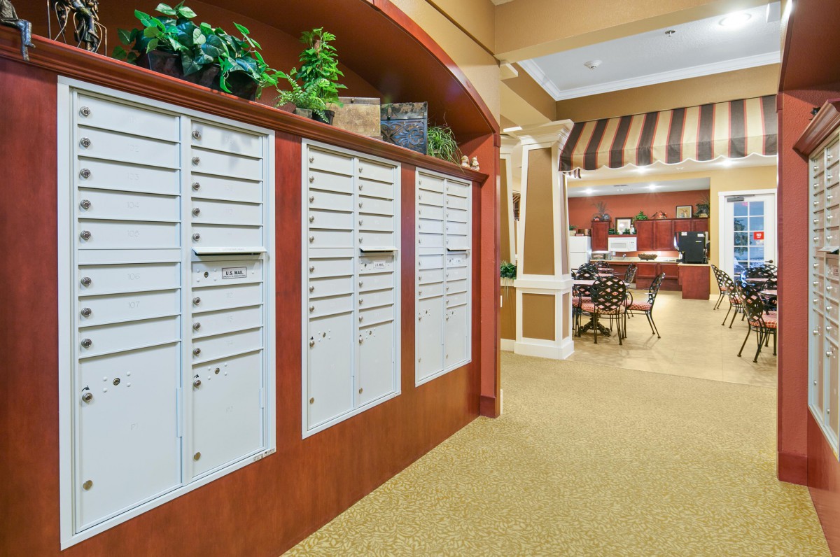 A lobby area featuring a row of mailboxes and a carpeted floor, with plants and an office space in the background.