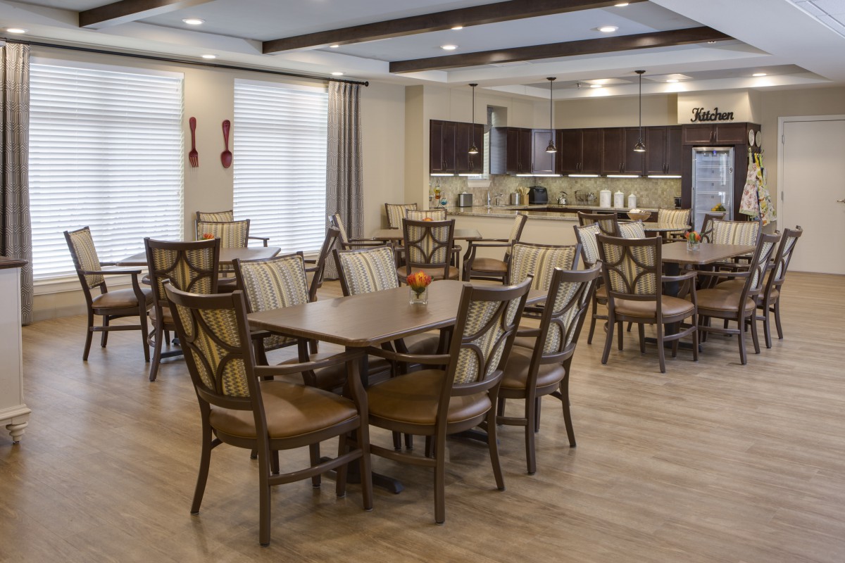 A modern dining room with multiple tables and chairs, featuring a kitchen area in the background.
