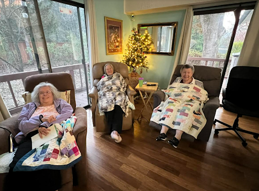 Three seniors enjoying a cozy afternoon by the window, wrapped in colorful quilts.