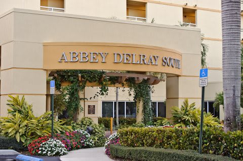 Entrance of a building with 'Abbey Delray South' sign, adorned with plants and flowers.
