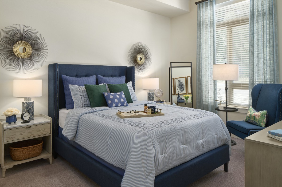 A cozy and well-decorated bedroom featuring a bed with multiple pillows, side lamps, a mirror, and a window with sheer curtains.