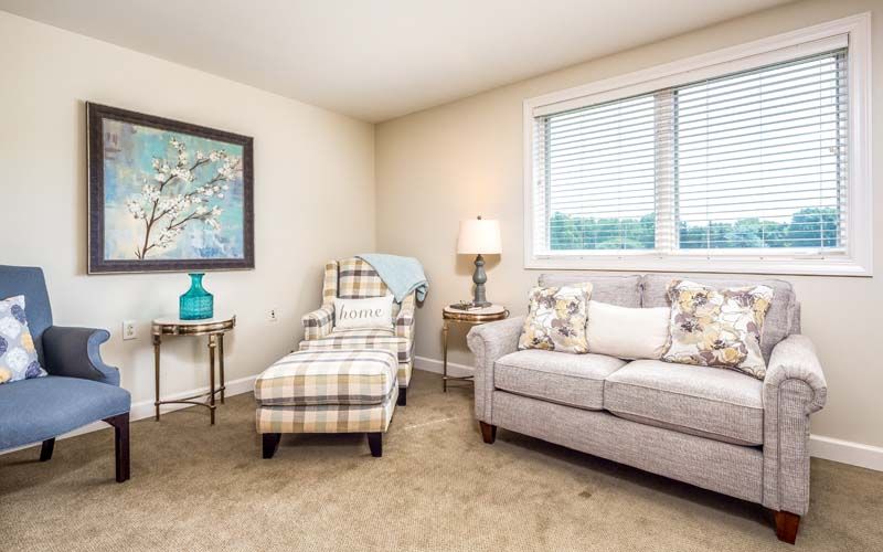 A cozy living room featuring a sofa, armchair with matching ottoman, side tables with decorative items, and a large window.