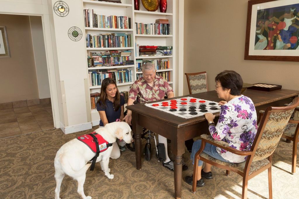A group of people playing checkers with a service dog nearby in a library setting.