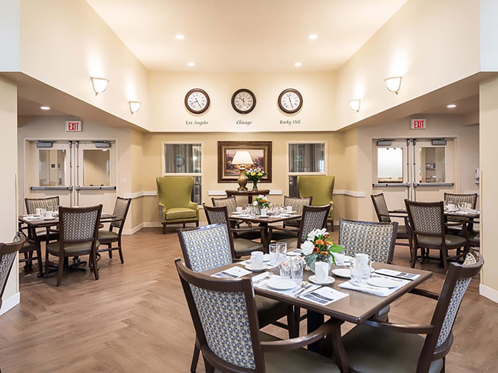 A well-lit dining room with neatly arranged tables and chairs, featuring elegant decor and multiple wall clocks.
