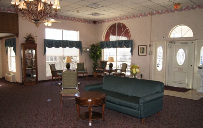 A cozy living room with green sofas and chairs, wooden tables, large windows, and decorative plants.