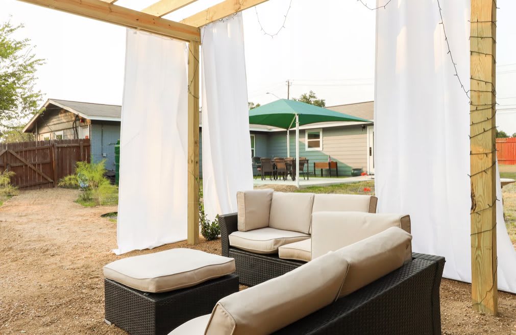 A cozy outdoor patio with comfortable sofa seating under a wooden pergola, complete with flowing curtains.