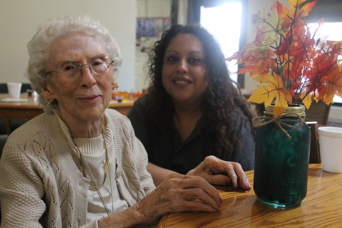 An elderly woman and a younger woman smiling together at a table with a vase of flowers.