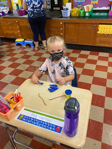 A child engaged in a crafting activity at a classroom table while wearing a mask.