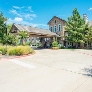 A spacious building entrance with a driveway, surrounded by trees and landscaping under a clear blue sky.
