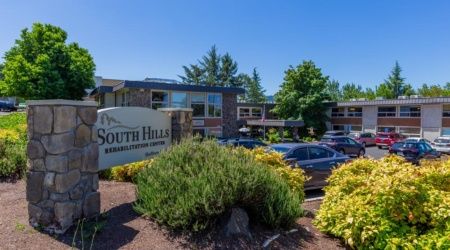 A commercial building with a sign reading 'South Hills,' surrounded by trees, shrubs, and a parking lot with cars.