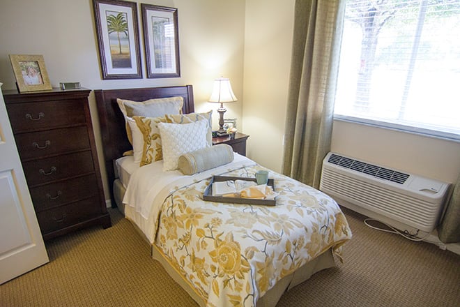 A cozy bedroom with a neatly made single bed, decorative pillows, and a bedside lamp.