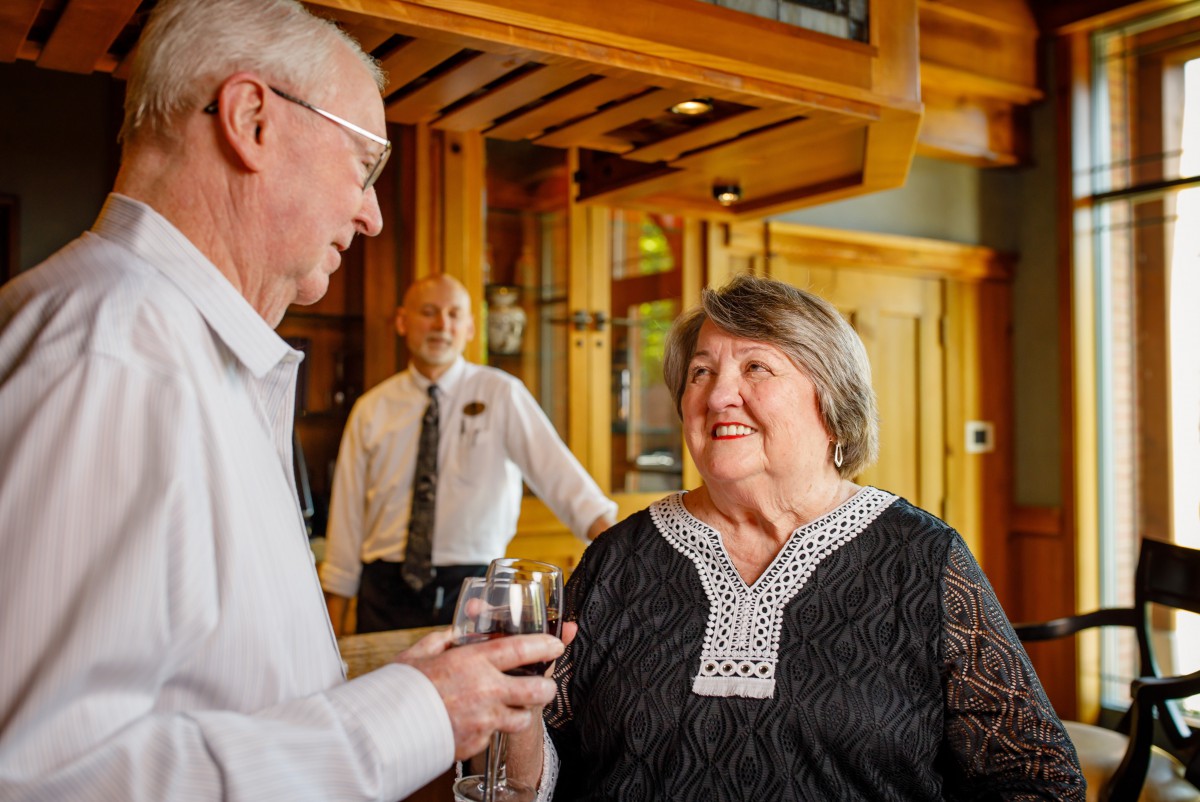 An elderly couple enjoys a conversation over wine in a restaurant, with a bartender in the background.