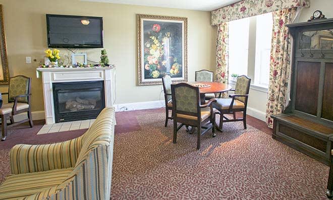 A cozy living room with a fireplace, table with chairs, and large windows adorned with curtains.