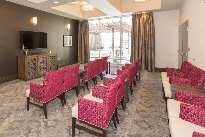 A modern conference room with rows of pink chairs facing a television, featuring large windows with curtains and patterned carpeting.