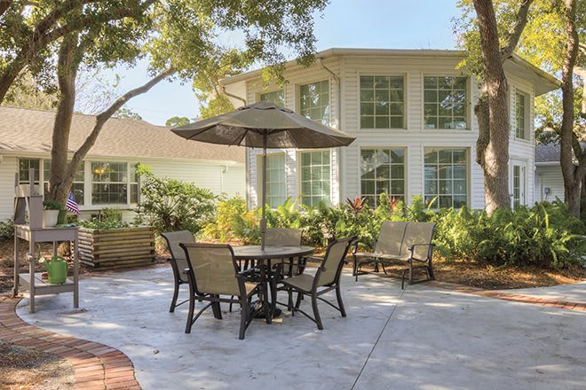 A cozy patio with outdoor furniture under a large umbrella, surrounded by lush greenery and adjacent to a residential building.