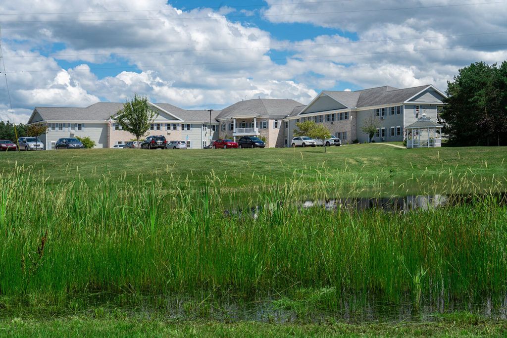 A suburban residential area with houses overlooking a grassy field and a small pond under a cloudy sky.