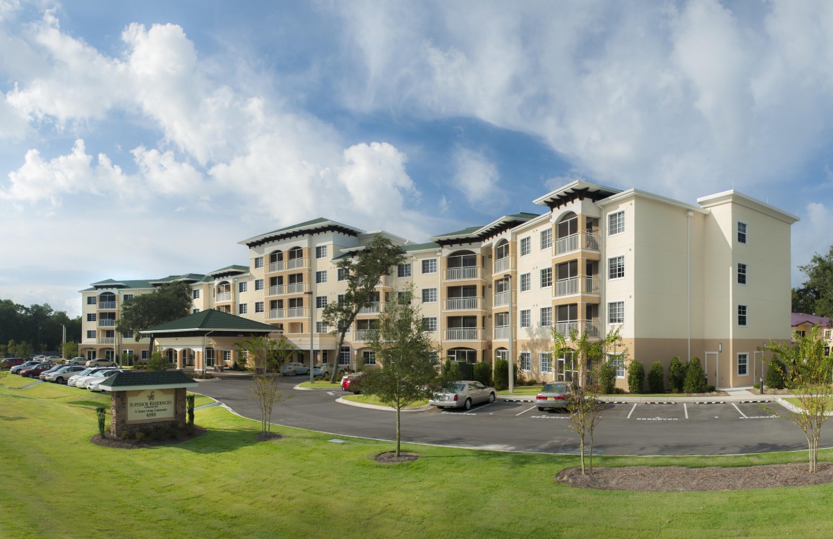 A modern apartment complex with a large parking lot and landscaped surroundings under a blue sky with clouds.