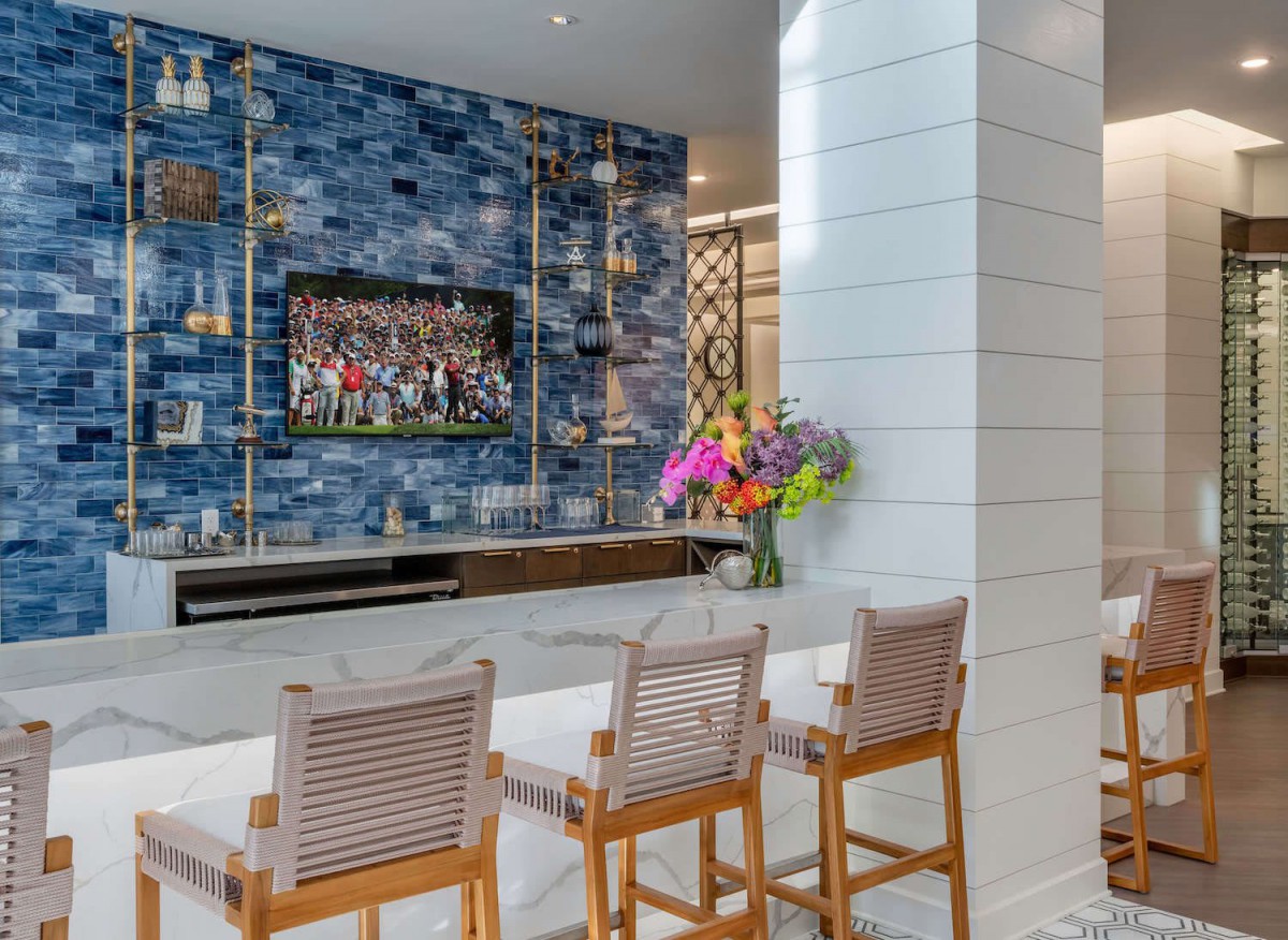 A modern living room featuring a blue tile accent wall with a mounted television, complemented by bar stools and a vibrant flower arrangement.