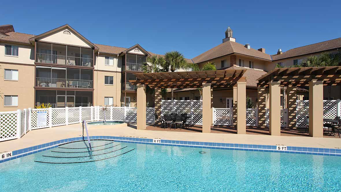 A sunny day view of an apartment complex with a swimming pool and pergola.