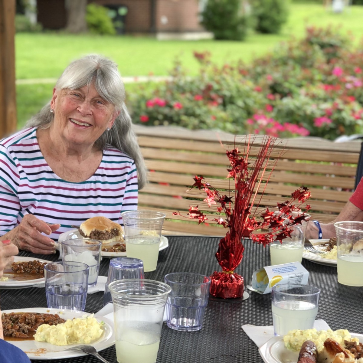 An elderly woman enjoying an outdoor picnic with food and drink under a gazebo in a garden.