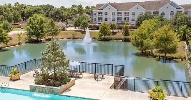A serene outdoor setting featuring a lake with a fountain, surrounded by trees and buildings, with a swimming pool in the foreground.