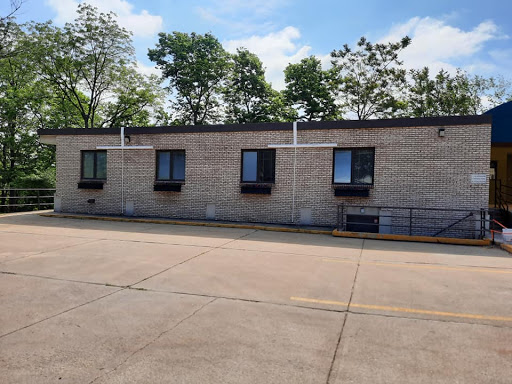 A view of a building with multiple windows, surrounded by trees and located next to a parking lot on a sunny day.