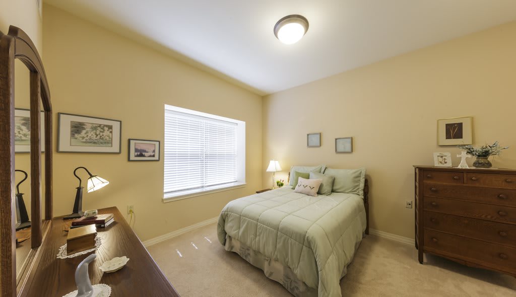 A cozy bedroom with a neatly made bed, soft lighting from a small lamp on the nightstand, and light streaming in through a window.